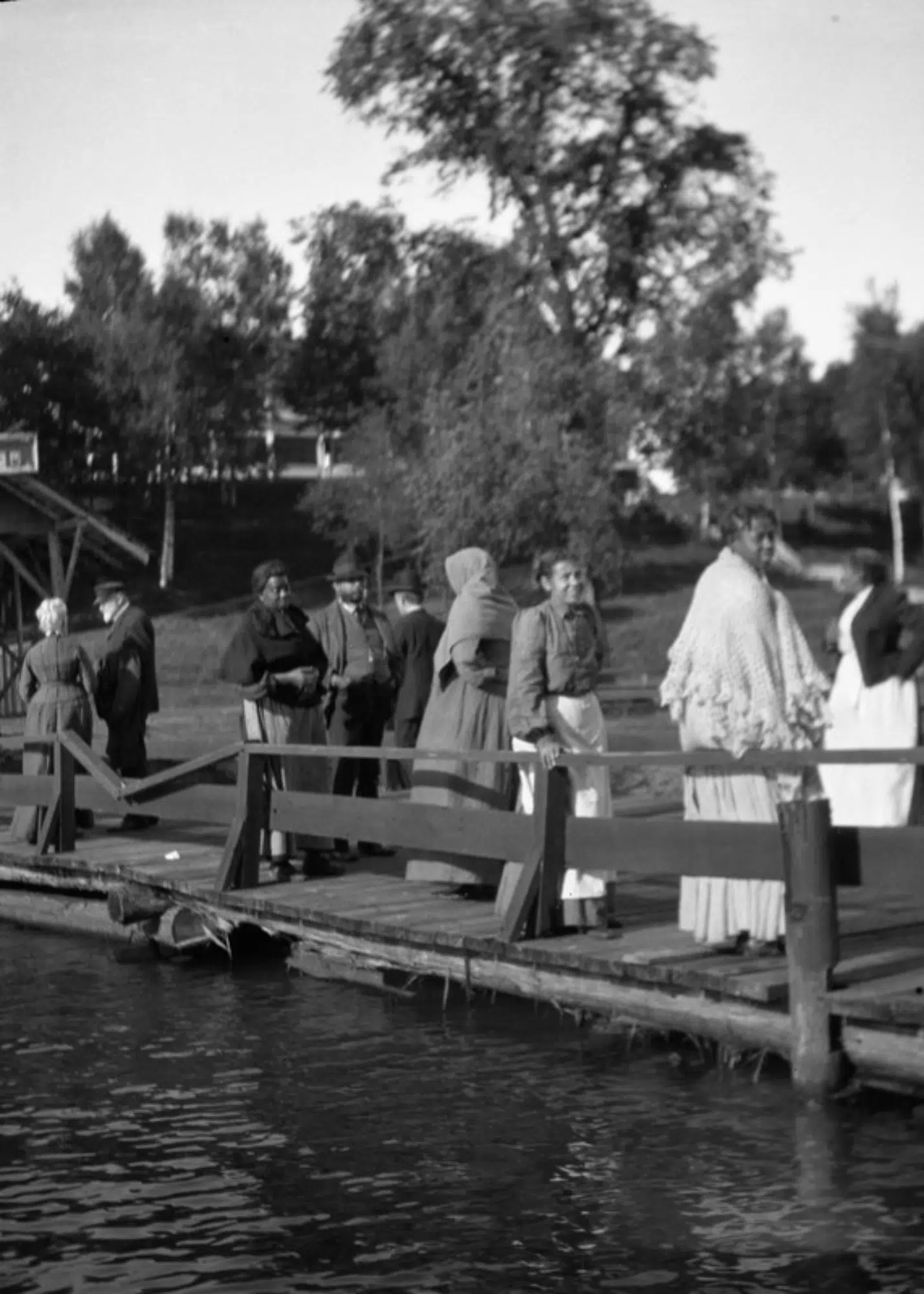 People on a dock at Les Cheneaux, probably landing at Les Cheneaux Club (note building in the background beyond the trees), ca. 1900-09.